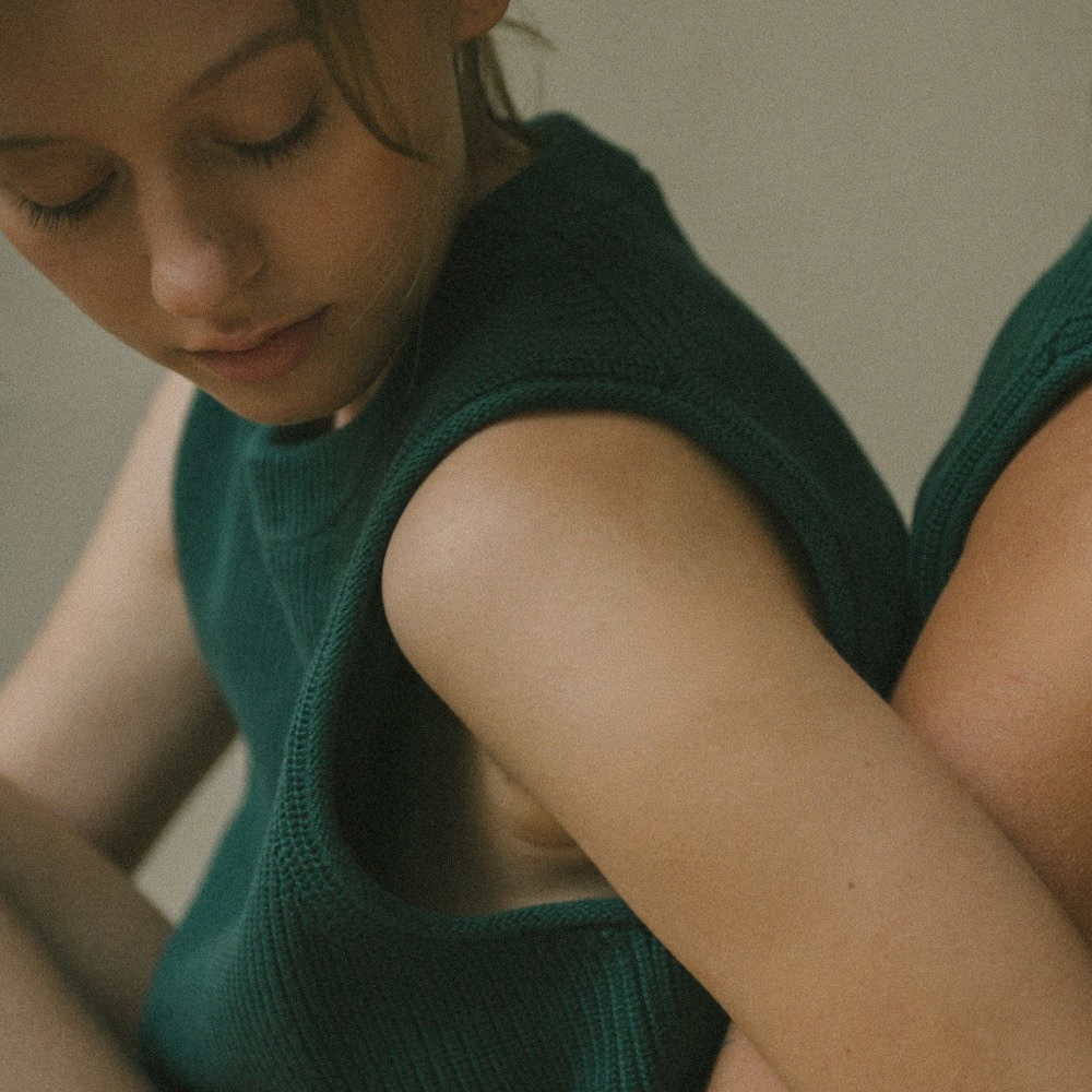 Jeune femme assise, genoux repliés contre elle, dans un haut en maille verte, le regard baissé